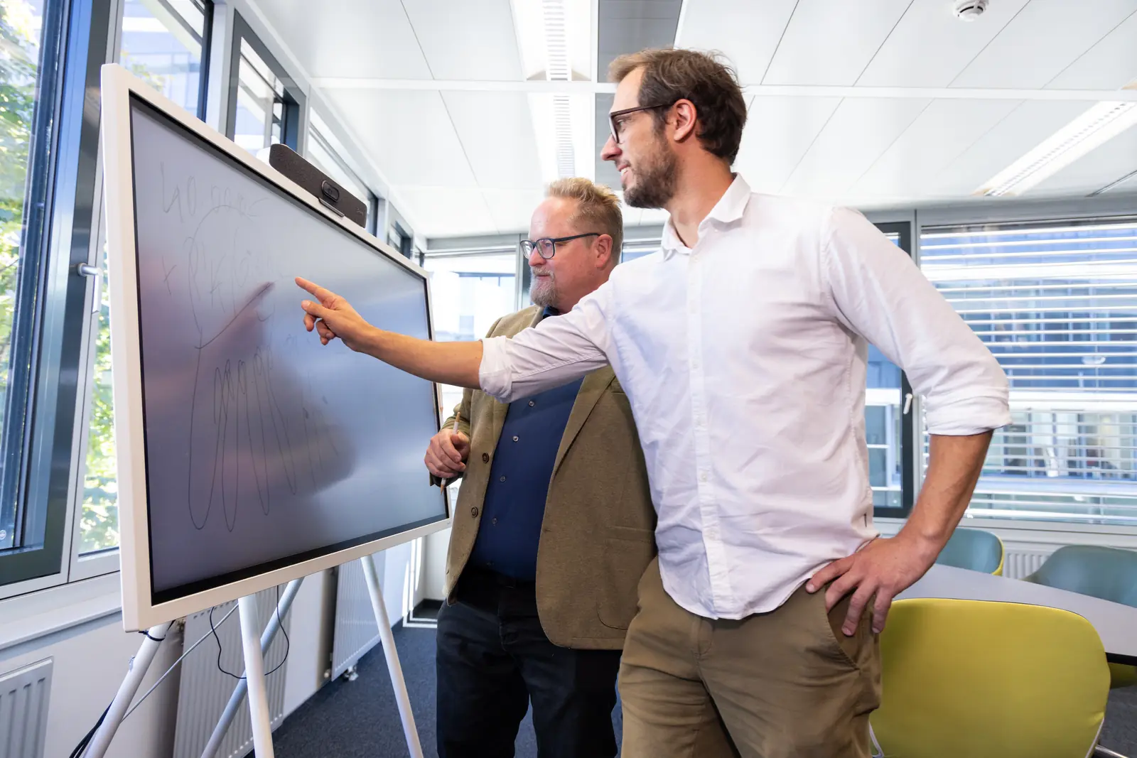 Consultant pointing at an overview on a large screen while a colleague listens