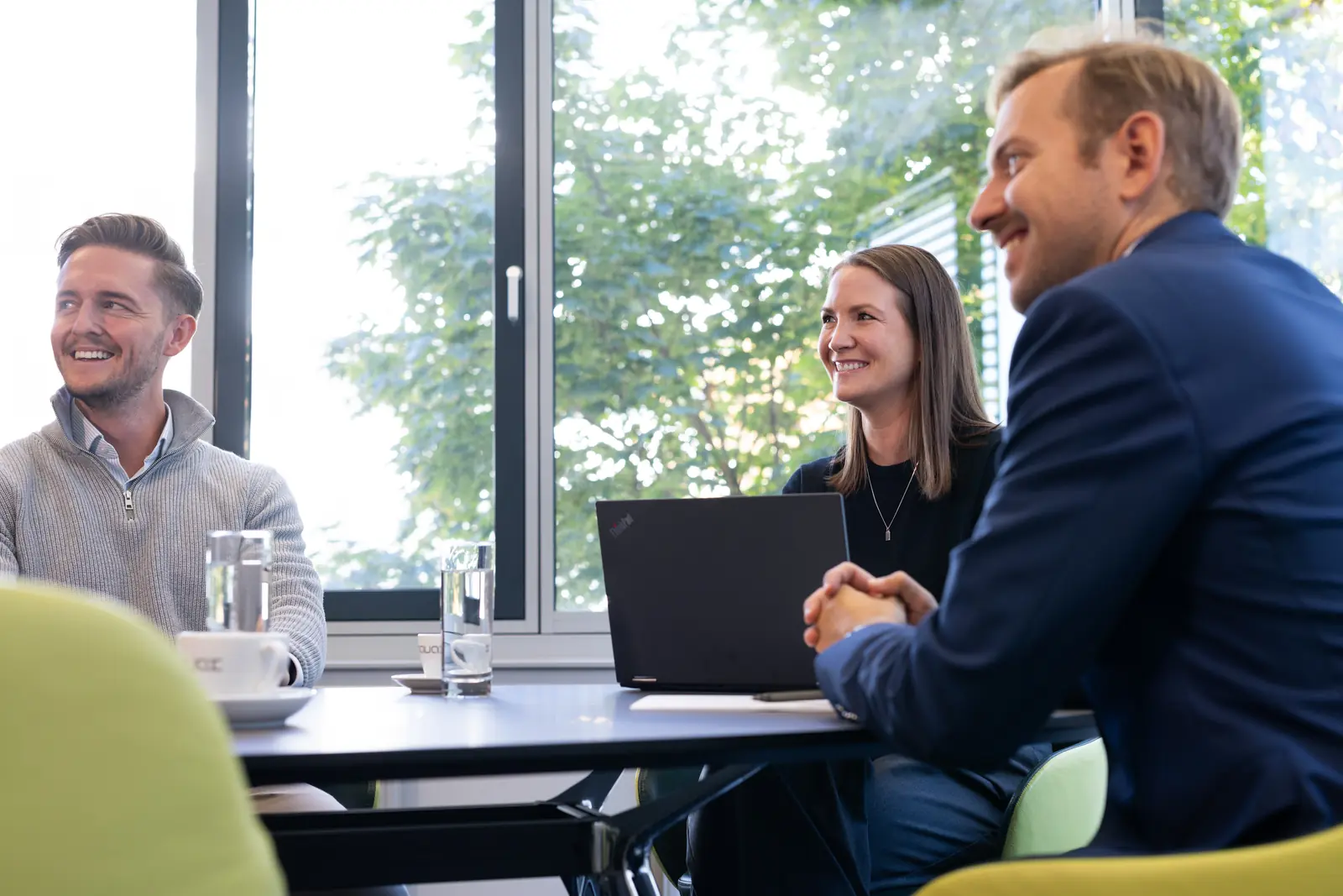 Drei Personen sitzen lächelnd an einem Besprechungstisch mit Laptop