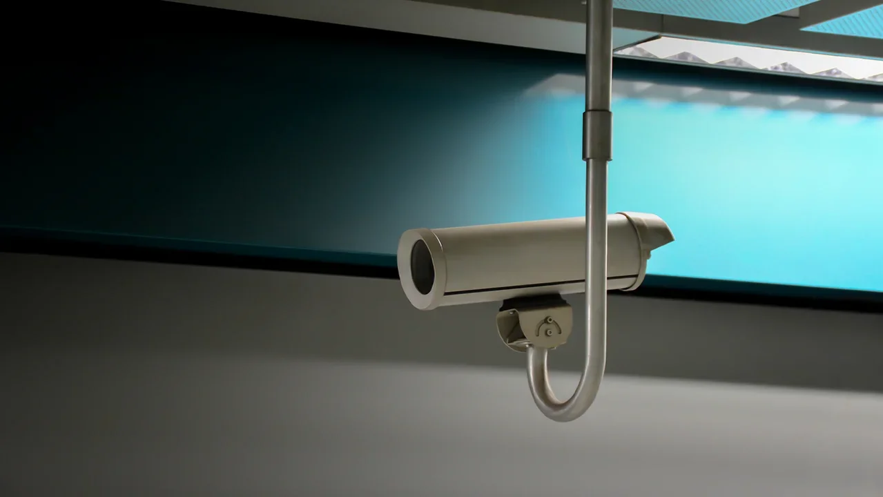 A surveillance camera on a ceiling mount in front of a lit wall