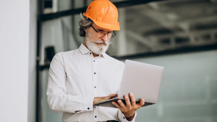 Mann trÀgt Bauhelm und hÀlt Laptop in der Hand